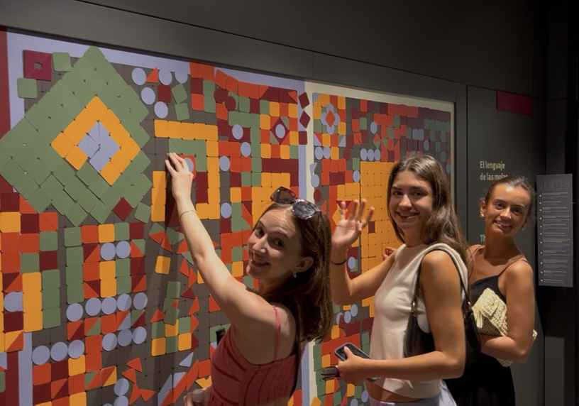 Three students smile while interacting with a colorful hands-on museum exhibit made of movable geometric shapes. Two students wave toward the camera as they rearrange pieces on the large interactive display.